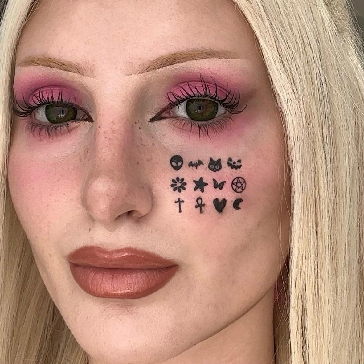 Close-up of a woman's face showing temporary black face decals applied to her cheek, including a skull, cat, bat, flower, star, pentagram, cross, ankh, heart, and crescent moon. She has blonde hair, pink eyeshadow, and brown lipstick.