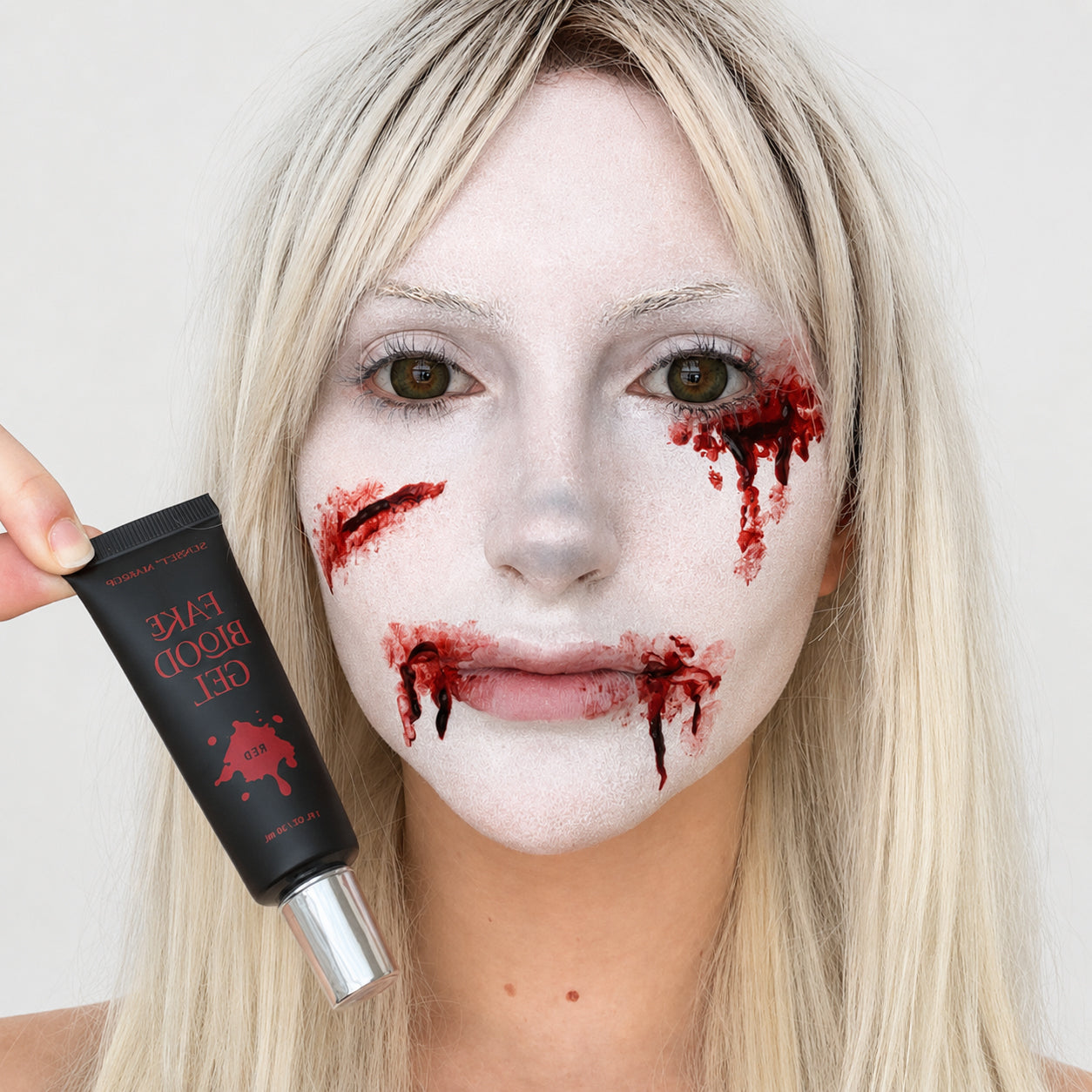 A blonde woman with white foundation face makeup and red fake blood splatter on her cheeks and mouth holds a black tube of Sunset Makeup Fake Blood Gel next to her face.