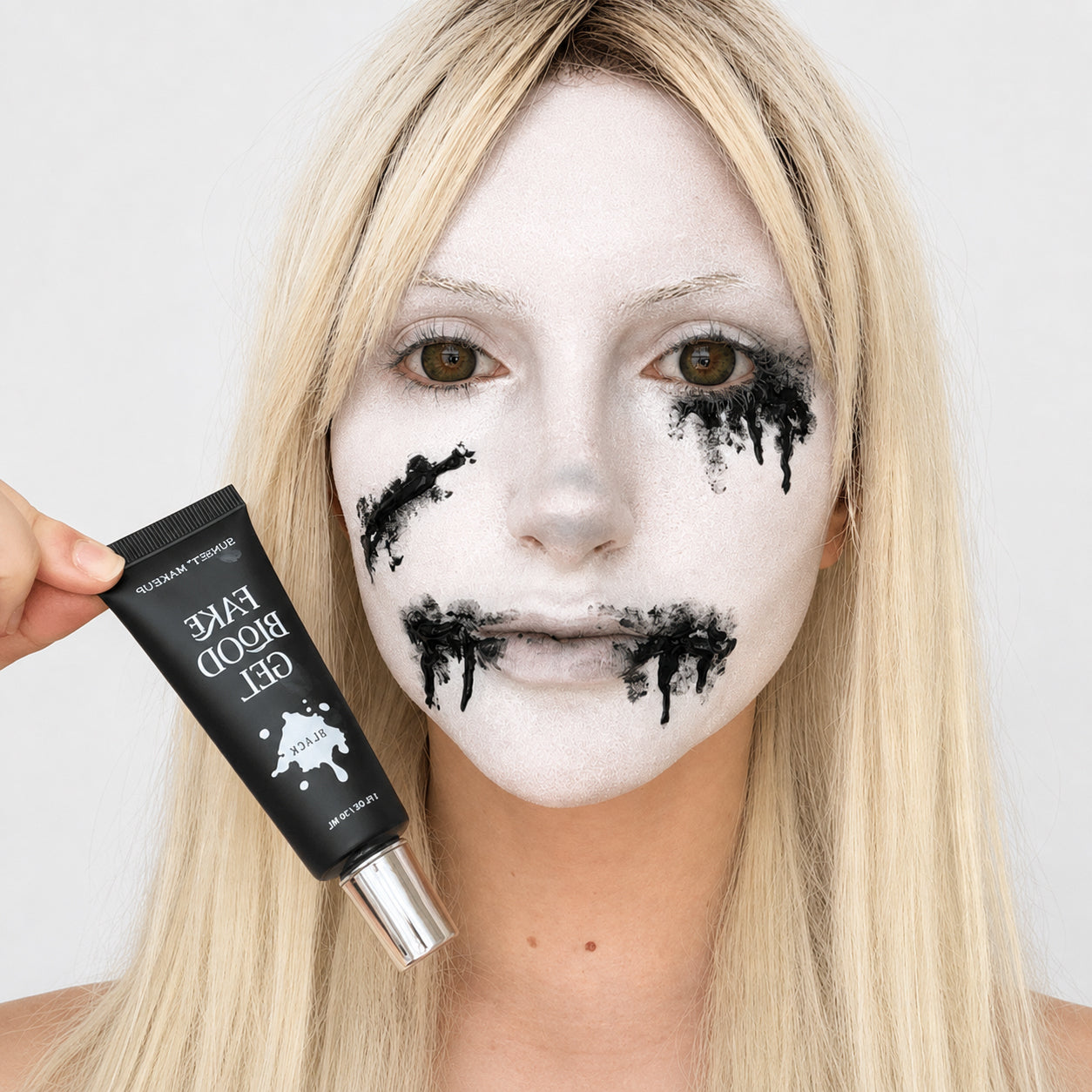 A blonde woman with white foundation face makeup and black fake blood splatter on her cheeks and mouth holds a black tube of Sunset Makeup Fake Blood Gel next to her face.