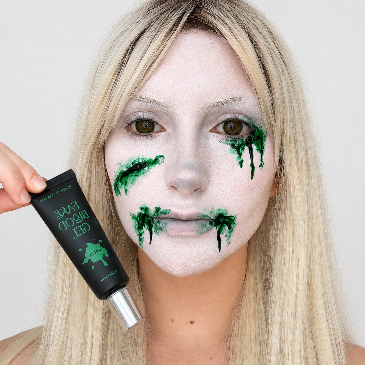 A blonde woman with white foundation face makeup and green fake blood splatter on her cheeks and mouth holds a black tube of Sunset Makeup Fake Blood Gel next to her face.