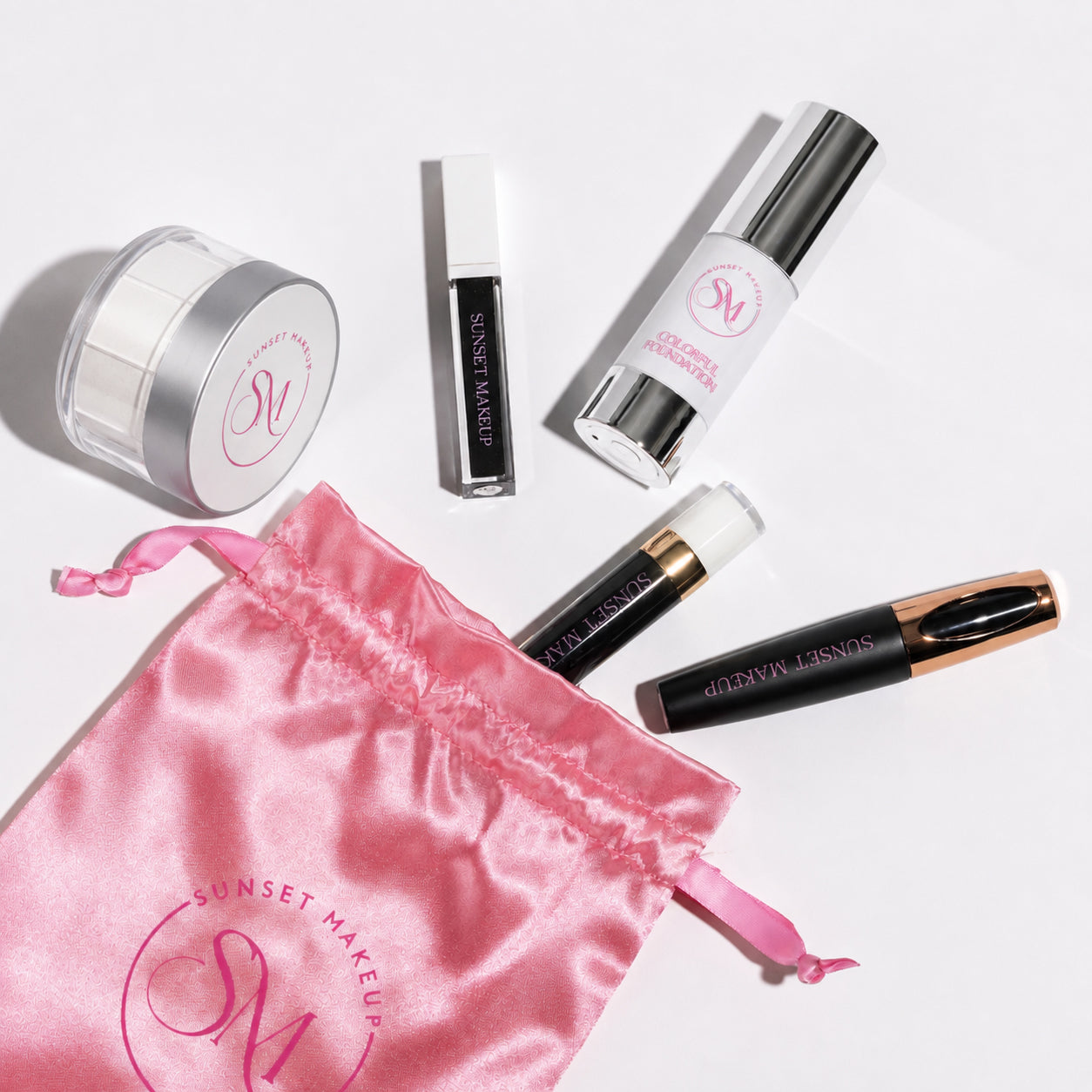 Sunset Makeup cosmetic set on a white background, products laying flat, including a pink satin drawstring bag, White Colorful Foundation, Black Matte Liquid Lipstick, White Mascara, Black Lip Gloss, and Setting Powder White.