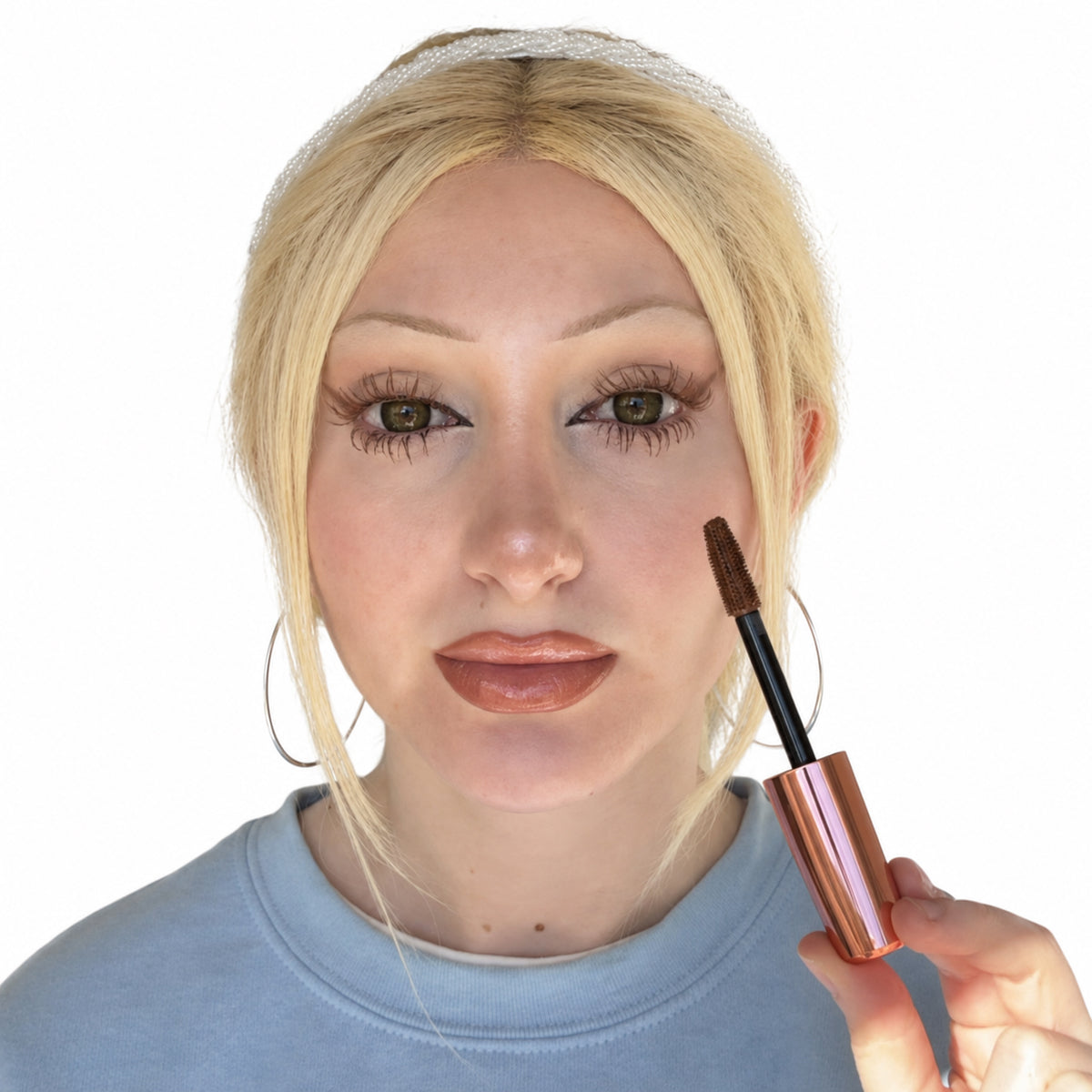 Model wearing light brown mascara on upper and lower lashes while holding the matching mascara wand against a white background.