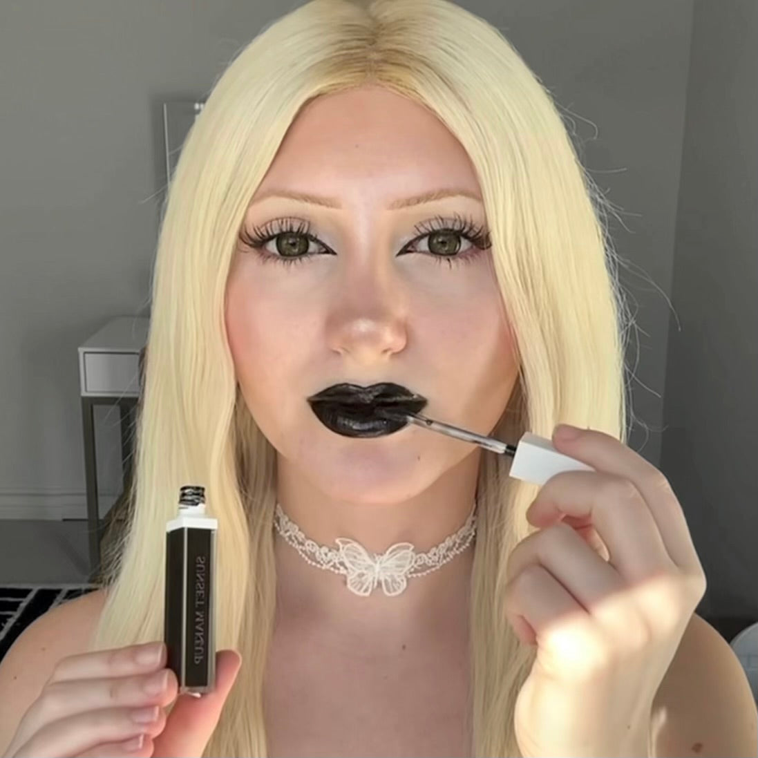 Sunset Makeup model with blonde hair applying black lip gloss to their lips, holding the tube of lip gloss in their left hand.
